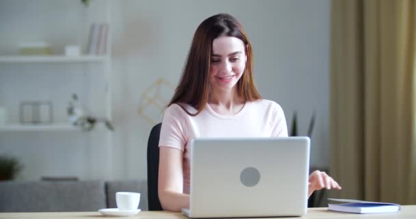 Joven mujer sonriente se sienta en casa en la mesa, mira a la pantalla de la computadora portátil moderna habla con el profesor en videollamada, estudio a distancia en e-learning en línea, mira el horario en el cuaderno lee notas, cuarentena — Vídeos de Stock