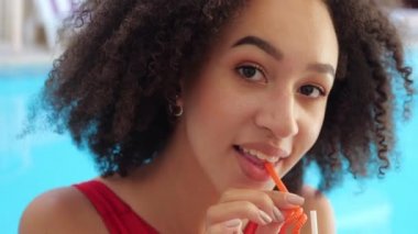 Afrika kökenli kıvırcık saçlı esmerin yakından çekilmiş portresi. Su havuzunun mavi zemininde oturmuş soğuk kokteyl mojito limonatası içiyor ve gizemli bir şekilde kameraya bakıyor.