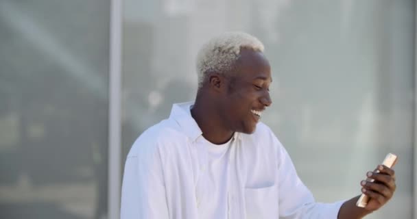 Schöner junger afrikanisch-amerikanischer Mann internationaler Student in modischer Kleidung, der in der Nähe einer städtischen Hauswand steht und mit einem Freund über eine Online-Videokonferenz mit dem Smartphone spricht — Stockvideo