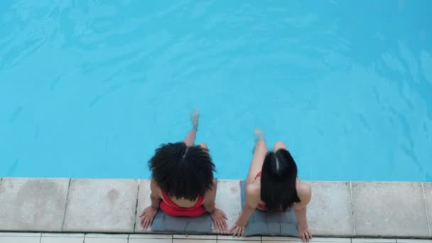 Two black haired girlfriends swing legs in water in public pool relax smile look at camera. African and chinese brunette students in red lingerie enjoy pastime in backyard together, view from above — Stock Video