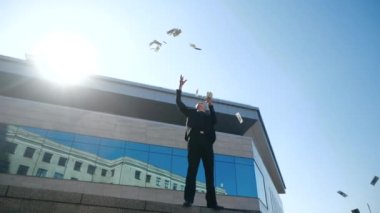 Başarılı genç iş adamı, kazanan açık havada mavi cam binanın yanında duruyor, şık bir siyah takım elbise giyiyor, havaya para atıyor, ikramiye kazanıyor, dolar dağıtıyor, başarı kavramı, alttaki görüş.