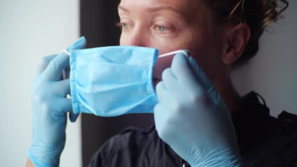 Jeune femme millénaire portant des gants de protection médicale met lentement masque protecteur jetable sur le visage. Médecin regarde par la fenêtre pendant la quarantaine auto-isolement COVID-19 pandémie en 2019-2020 ans — Video