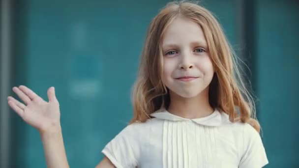 Portrait of cute little girl with long golden blonde hair and blue eyes, standing looking at camera, waves his palm, makes welcome gesture, sign of hello. Close up child moving hand in goodbye motion — Stock Video