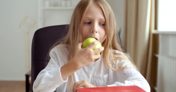 Портрет школьницы сидит за столом в домашней школе, улыбается, берет зеленое яблоко, кусает и ест фрукты, наслаждается здоровой пищей с витаминами, чувствует голод во время перерыва, восстанавливает силы во время обеда — стоковое видео