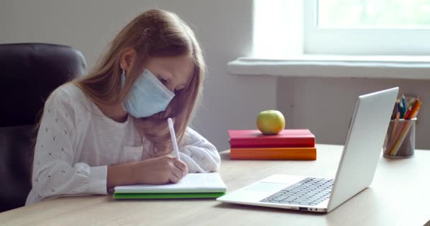 Chica de la escuela en los estudios de máscara protectora en casa con el profesor de Internet. Niño mira a la computadora portátil, escribir en el cuaderno, educación en línea, video llamada tutoría, lección de la escuela a distancia en cuarentena covid-19 — Vídeos de Stock