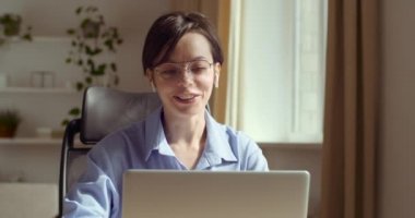 Gözlüklü genç bir kadın kulaklık takıyor web kamerasına konuşuyor uzaktan video konferans görüşmesi yapıyor ve dizüstü bilgisayarda yazıyor. Kadın internet öğretmeni evde çene çalıyor, tele çalışma kavramı var.