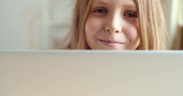 Lindo colegial se esconde detrás de la computadora portátil con la cara sonriente.Cierre los ojos femeninos mirando desde la pantalla del ordenador. Retrato de la niña adolescente divertida jugar en la mesa haciendo la tarea en línea en la red, la infancia feliz — Vídeo de stock