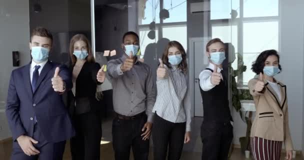 Multi etnikai csapat fiatal ambiciózus alkalmazottak visel arc maszk, pózol pozitív hüvelykujj fel gesztus, mutatnak támogatást, mint jel, igen szimbólum, áll az irodában karantén világjárvány covid vírus — Stock videók