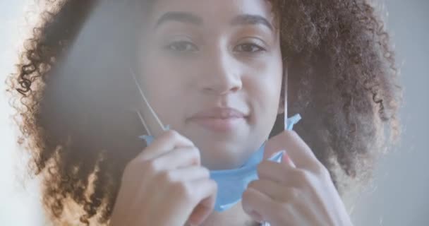 カメラを見て女性の顔を閉じます。アフリカ系アメリカ人女性医師看護師の肖像は、太陽光の下でポーズ鼻と口の青の医療保護マスク、 covidウイルスパンデミックの概念を置きます — ストック動画