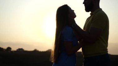 Genç ve mutlu Avrupalı çifti öpmek için romantik bir tarih. Dışarıda, yaz günbatımının arkaplanına karşı yavaş çekimde. Saati olan sakallı adamın yakından görüntüsü. Güneşin altında güzel bir kız arkadaşa sarılıyor..