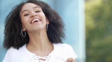Neşeli, güzel, kıvırcık, kıvırcık, Afro-Amerikan genç bir kadın akıllı telefonuyla atlamayı, heyecan verici duygularından kollarını sallamayı, modern şehir caddesinde neşeyle gülmeyi ve mutlulukla gülümsemeyi seviyor..
