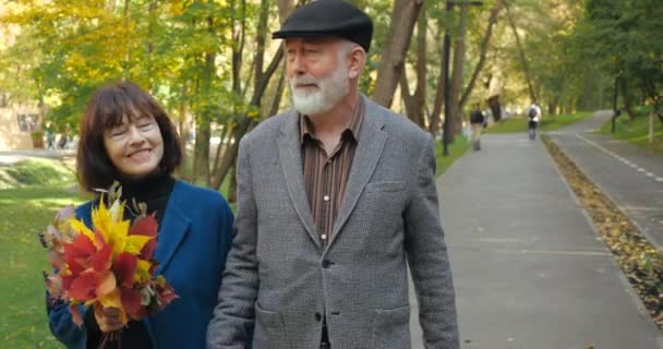 Atraktivní usměvavý starší pár vousatý manžel a manželka s kyticí podzimních listů si vychutnat aktivní procházku v parku. Šťastní prarodiče se procházejí venku na čerstvém vzduchu, poblíž předního výhledu. — Stock video