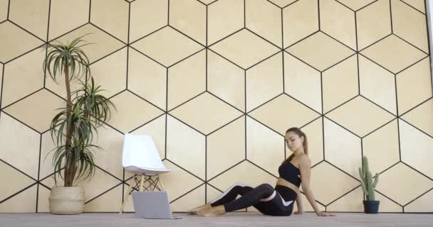 Jeune athlète danseuse femme millénariste siège sur le sol à la maison dans la chambre, regarde l'écran d'ordinateur portable de l'appareil, répète des exercices sportifs en ligne en réseau, fait des exercices d'aérobic, trains jambes avec bande élastique — Video