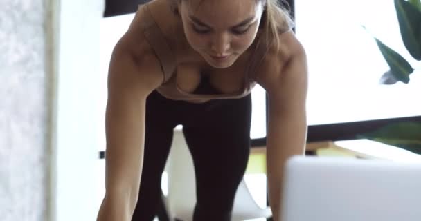 Молода сильна жінка займається спортом вдома з відео уроку на ноутбуці онлайн крупним планом. Тисячолітня дівчина в спортивному одязі робить фізичні вправи на тілі відсутні на підлозі, стоячи в позі, ізольований карантин — стокове відео