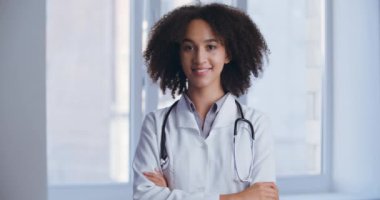 Klinikte durmuş, kollarını kavuşturup kameraya bakarken gülümseyen genç, etnik hemşire kadın. Olumlu insan cerrahisi kadın memnuniyet ve neşe hissediyor, dost canlısı tıbbi profesyonel