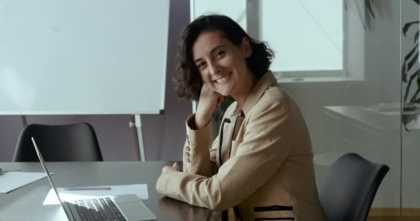 Πορτρέτο μιας νεαρής χαμογελαστής γυναίκας επιχειρηματία, χαρούμενη μαθήτρια, φιλική δασκάλα που φοράει σακάκι, κάθεται στο τραπέζι μπροστά από το laptop, δουλεύει online, ξεκουράζεται στο διάλειμμα, κοιτάζει την κάμερα χαρούμενα, πλαϊνή άποψη — Αρχείο Βίντεο