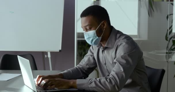 Νεαρός Αφρικανός με αυτοπεποίθηση επιχειρηματίας αλλοδαπός φοιτητής με ιατρική προστατευτική μάσκα δακτυλογράφησης σε φορητό υπολογιστή, που εργάζεται εξ αποστάσεως σε απευθείας σύνδεση κατά τη διάρκεια πανδημίας ιού, κρατώντας σημειώσεις στο σημειωματάριο, κάνοντας την εργασία στο διαδίκτυο — Αρχείο Βίντεο