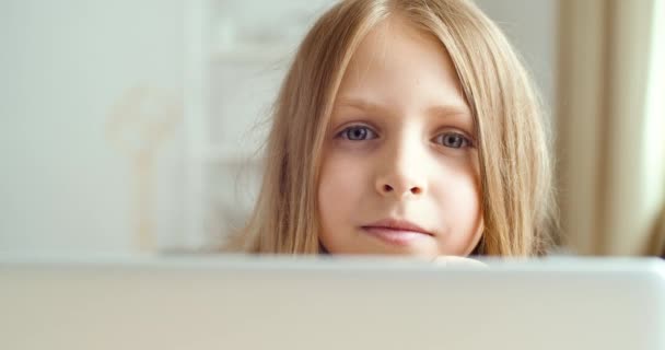 Ritratto di bambina bionda seduta al computer portatile a casa o in classe, bambino guarda la macchina fotografica con gli occhi azzurri, sorride carino, fa i compiti, studia online, e-learning concetto in quarantena — Video Stock