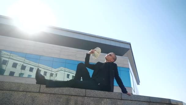 Proud rich man lies on tile against background of business center and blue sky, holds stack of dollars in his hands, makes fan of money banknotes, brags about prize or win, concept of successful deal — Stock Video