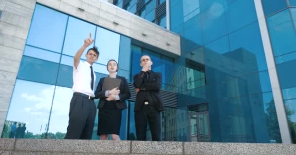 Successful business people discuss business and stand outdoors. Two young men and one woman in suit communicate and walk near office building of business center, negotiate, discuss project ideas deal — Stock Video