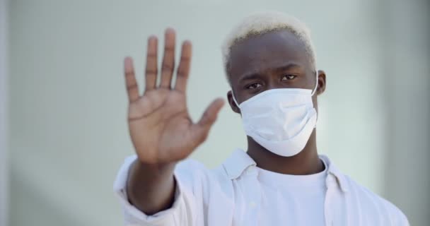 Joven chico negro étnico lleva máscara en la cara, se protege del virus, muestra señal de stop con la mano, pone la palma hacia adelante, prohibiéndole acercarse, hace frontera de distancia, peligro de infección con el virus — Vídeos de Stock