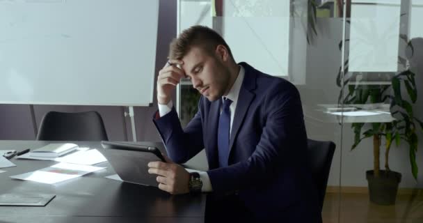 Un homme d'affaires confiant et concentré porte un costume, est assis à table au bureau, utilise une tablette numérique pour travailler en réseau, regarde l'écran de l'appareil, lit les nouvelles en ligne, écrit des notes dans un carnet, planifie un projet futur — Video