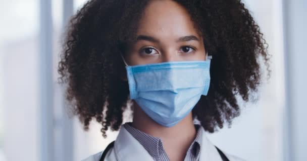 Close-up van vrouwelijk gezicht in medisch beschermend masker, jonge Afro-Amerikaanse vrouw student arts verpleegkundige staat binnen serieus kijken naar camera, tijd van hevige virus pandemie, kliniek ziekenhuis concept — Stockvideo