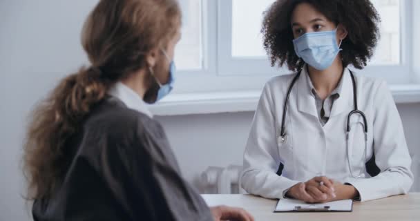 Male patient caucasian millennial with long hair, sits at reception of young woman doctor, wears protective mask, suffers from cough disease, feels soreness, needs medical help, view from behind — Stock Video