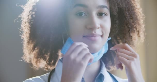 Närbild porträtt av afroamerikanska lockigt hår kvinnlig läkare sätter på medicinsk skyddsmask i modern klinik eller sjukhus. Stoppa viruset. Ung sjuksköterska stående poserar, allvarligt titta på kameran — Stockvideo