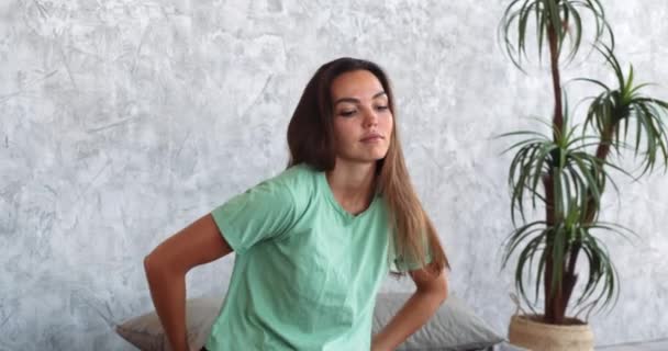 Beautiful brunette woman wears green T-shirt, does exercises in morning while sitting in soft bed in bedroom at home or in hotel room, stretches muscles of body, start of active day, lazy workouts — Stock Video
