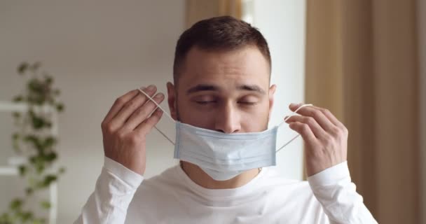 Porträtt av ung stilig man sätta medicinsk mask på ansiktet som skydd respirator från sjukdom som coronavirus. Man tittar på kameran, står hemma eller på kontoret inomhus ensam. COVID-19-konceptet — Stockvideo