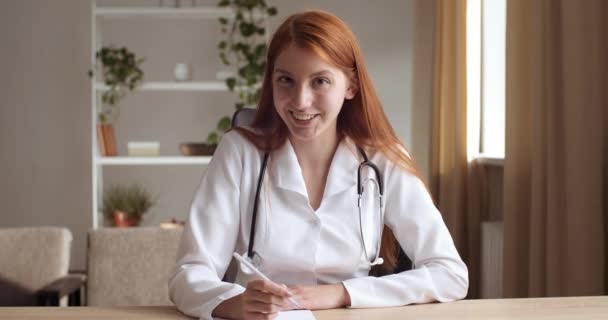 Młoda ruda profesjonalna kobieta lekarz nosić mundur ze stetoskopem rozmawiać przez telekonferencję rozmowy, skonsultować się z pacjentem online patrząc na kamerę internetową, pisać w notebooku, odległe konsultacje lekarskie — Wideo stockowe