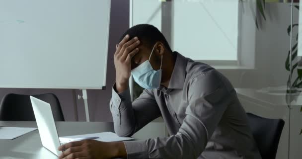 動揺若いアフリカのビジネスマンは、ラップトップの前に座って、医療マスクを身に着けている、 covidウイルスの隔離発生の継続についてのオンライン悪いニュースを読み取り、頭痛やストレスを感じ、損失の概念 — ストック動画