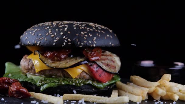 Zbliżenie duży soczysty czarny burger ze smażonymi frytkami leży na pokładzie lub stół w kuchni w kawiarni bar tłusty serdeczny hamburger niezdrowe fast food, sól spada na cheeseburgera z góry — Wideo stockowe
