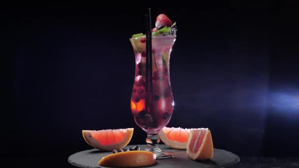 Zbliżenie szklanki z koktajlem owocowym, jagody pływają w wodzie alkoholowy napój alkoholowy ze słomką, truskawki i kawałek ogórka stoi na stole kuchennym baru otoczony plastrami grejpfruta — Wideo stockowe