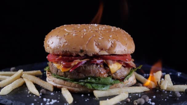 Burger besar lezat terletak pada panggangan hitam, hamburger dengan sayuran dan daging sapi berlubang digoreng pada barbekyu di atas api, junk makanan cepat saji Burger keju dan kentang goreng yang tersebar di permukaan — Stok Video