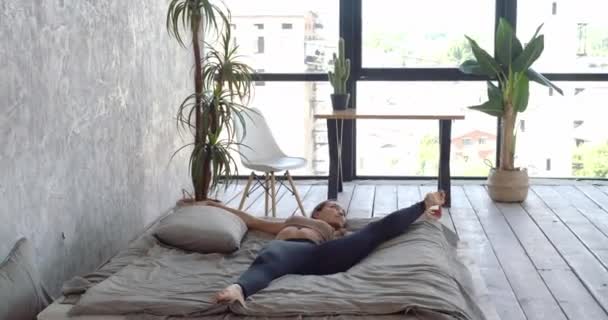 Движущаяся рамка молодая женщина в спортивной одежде лежит на мягкой удобной стильной кровати в спальне на фоне современных столов и комнатных растений, поднимает ногу, тренирует мышцы, делает упражнения на растяжку — стоковое видео