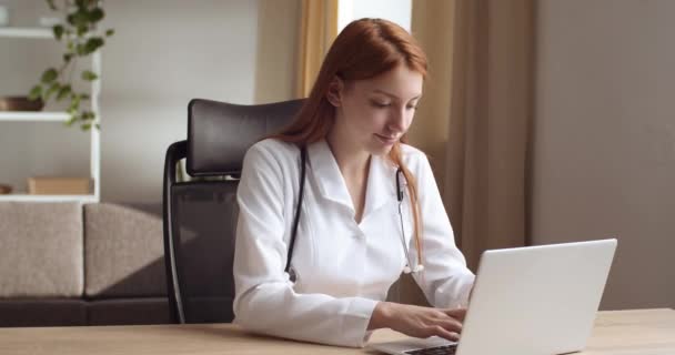 Ernste rothaarige junge Ärztin oder Krankenschwester sitzt mit Laptop im Büro der Klinik, liest gute medizinische Nachrichten, schreit freudig, fühlt Erfolg und Glück und feiert den Sieg — Stockvideo