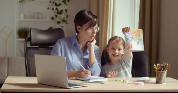 Pěkná malá holčička sedící s matkou nebo učitelkou u stolu doma nebo ve třídě, kreslí na papíře, představuje pestrobarevný obrázek, pořady před kamerou. Starší sestra pomáhat dítě se studiem, bavte se s dětmi — Stock video