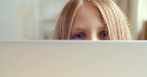 Portret małej uroczej dziewczynki chowającej się za laptopem podczas siedzenia przy stole. Zbliżenie kobiet niebieskie oczy dziecka patrząc zza ekranu komputera, bawi się podczas zajęć, pozwala na odrabianie lekcji — Wideo stockowe