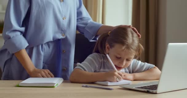 Mam controleert huiswerk van de kleuter dochter, meisje zit thuis tafel schrijft op papier, vrouw strijkt kind op hoofd goedkeuring les van haar jongere zus, leraar prijst student, concept van e-learning — Stockvideo