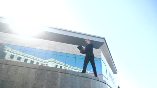 Νέος όμορφος επιχειρηματίας φορώντας μαύρο κοστούμι, περπατώντας στο κέντρο της πόλης με έγγραφα στο φάκελο στο χέρι, διαβάζει σημειώσεις για να πάει, ελέγχει σύμβαση κινείται με αυτοπεποίθηση βάδισμα σε φόντο του κτιρίου — Αρχείο Βίντεο