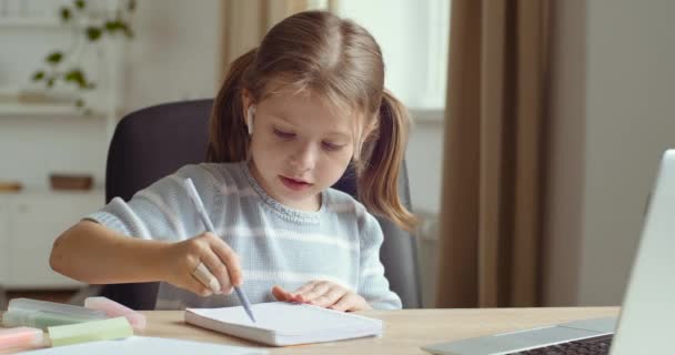 Küçük sevimli anaokulu kızının portresi kulaklık takıyor evde tek başına oturuyor dizüstü bilgisayarın hareketlerine bakıyor internetteki öğretmenlerin kağıt üzerine kalem ve uzaktan öğrenim konseptiyle hareketlerini tekrar ediyor. — Stok video