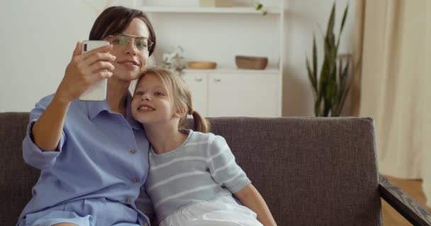 Giovane madre amorevole con la sua piccola figlia prescolastica seduta su un morbido divano confortevole a casa, scattando selfie al telefono. Donna e bambina scattano foto o video sulla fotocamera dello smartphone insieme sorridendo — Video Stock