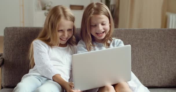Två små flickor sitter på soffan tittar på laptop, blonda systrar hemma tillsammans titta på film eller video på nätet i datorskärmen, par flickor vänner tillbringa tid tillsammans skratta ha kul — Stockvideo