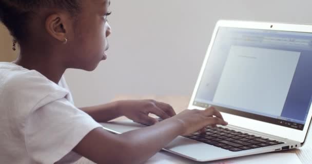 Боковой вид маленькой школьницы смешанной расы африканского происхождения, сидящей в домашней школе за ноутбуком, играющей на клавиатуре, пишущей историю онлайн, делающей домашнюю работу — стоковое видео