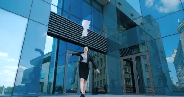 Gelukkige jonge zakenvrouw viert succes, komt uit het moderne kantoor, gooit documenten in de lucht. Aantrekkelijke vrouwelijke ondernemer in zwart pak wandelen in de buurt van modern gebouw, strooit papieren weg — Stockvideo