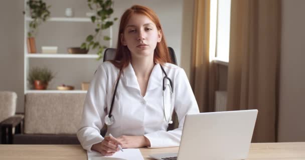 若い女性の医師は、医療の制服を着て、ノートパソコンで事務所の診療所に座って、窓の外を見て、カメラに彼の視線を回し、彼の手の中に優しい保持ペン笑顔。看護師像 — ストック動画