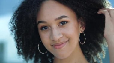 Kendine güvenen güzel bir Afrikalı kız gülüyor. Mutlu etnik Amerikalı öğrenci, kıvırcık saçlı, yuvarlak küpeli, hafif makyajlı ve çilli gülümsemeli ve sağlıklı beyaz dişlerini gösteriyor