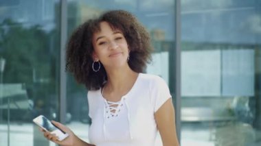Neşeli genç Afro-Amerikan milenyum kadını yükseğe zıplamaktan, silah sallamaktan ve elinde akıllı telefonuyla havada dans etmekten, modern şehir binasının arka planına gülüp eğlenmekten hoşlanıyor..